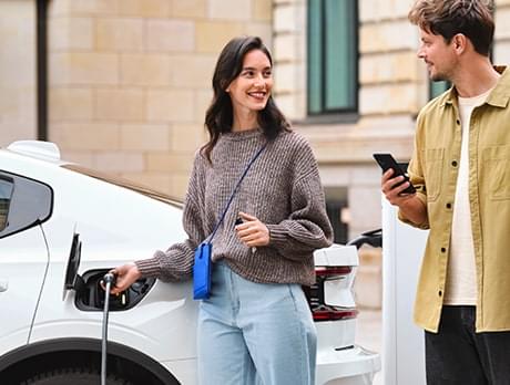 Woman stands next to an electric vehicle, looking at the CHARGE NOW app.